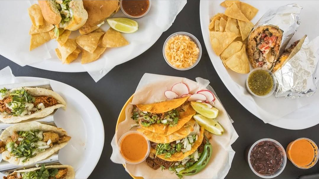 An overhead spread of Maria's Tacos plates: griddled birria-style tacos, street tacos, a torta and a burrito with chips and salsas
